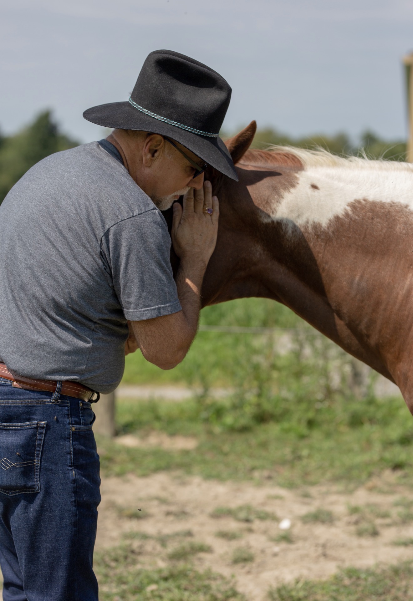 Man in a cowboy hat petting a brown and white horse on the ground surrounded by trees